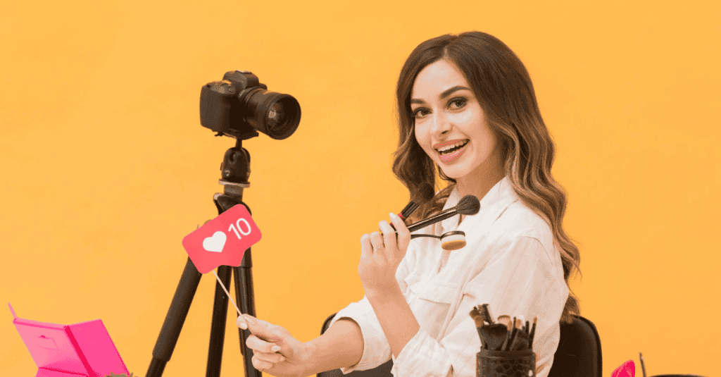 A smiling content creator sits in front of a camera, holding makeup brushes and a social media "like" sign, ready to film a beauty tutorial.