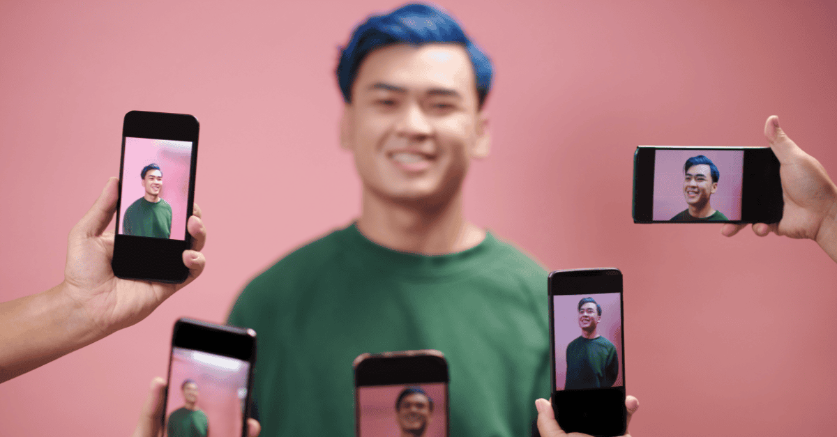 A smiling man with blue hair poses in front of a pink background while multiple people capture his photo on smartphones, symbolizing influencer collaboration or social media promotion.