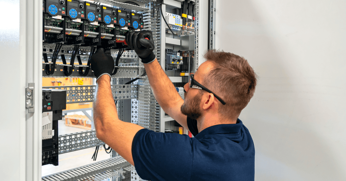 An electrician wearing safety glasses and gloves works on wiring inside an electrical control panel, adjusting components with a screwdriver. 