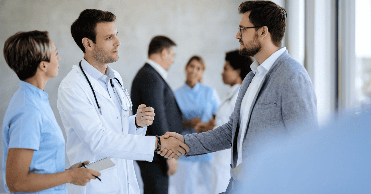 A doctor in a white coat shakes hands with a professionally dressed man in an office setting, with medical staff and other professionals conversing in the background.